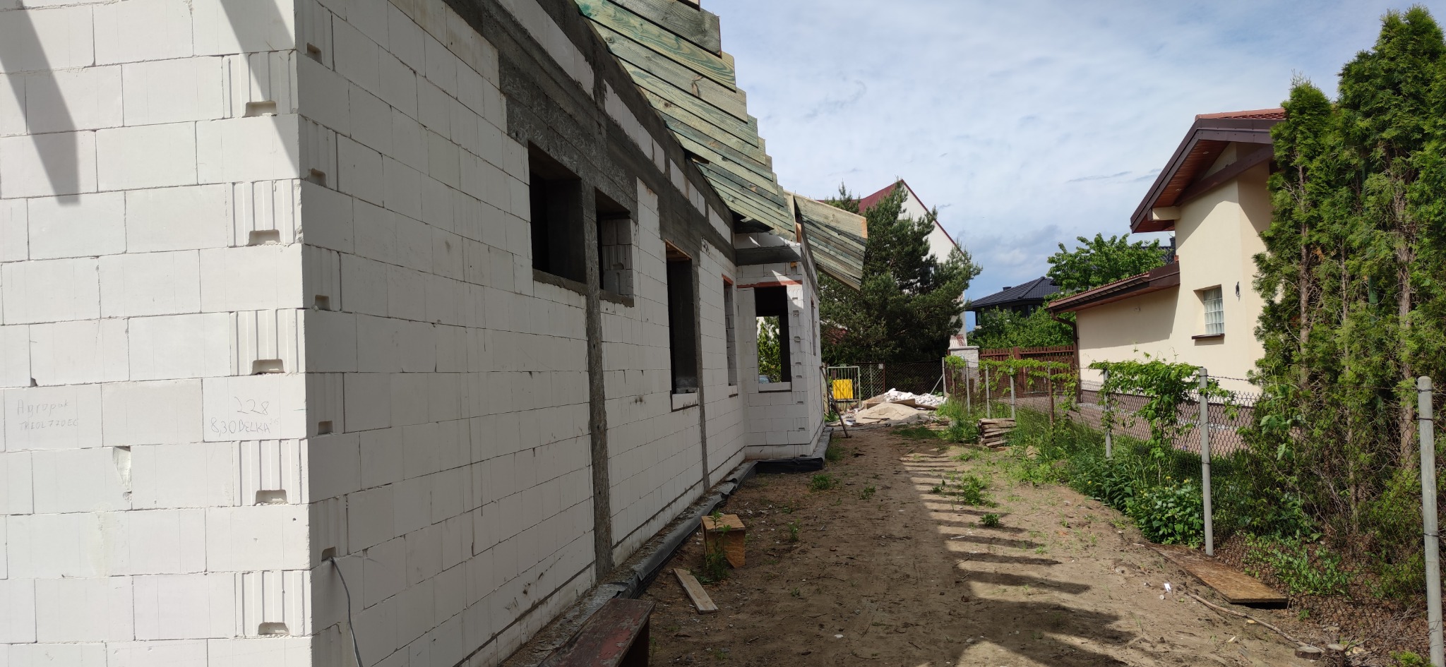 Budowa domu jednorodzinnego z białych bloczków, widoczne otwory okienne i przygotowane elementy więźby dachowej, na ścianach widoczne napisy producenta bloczków.