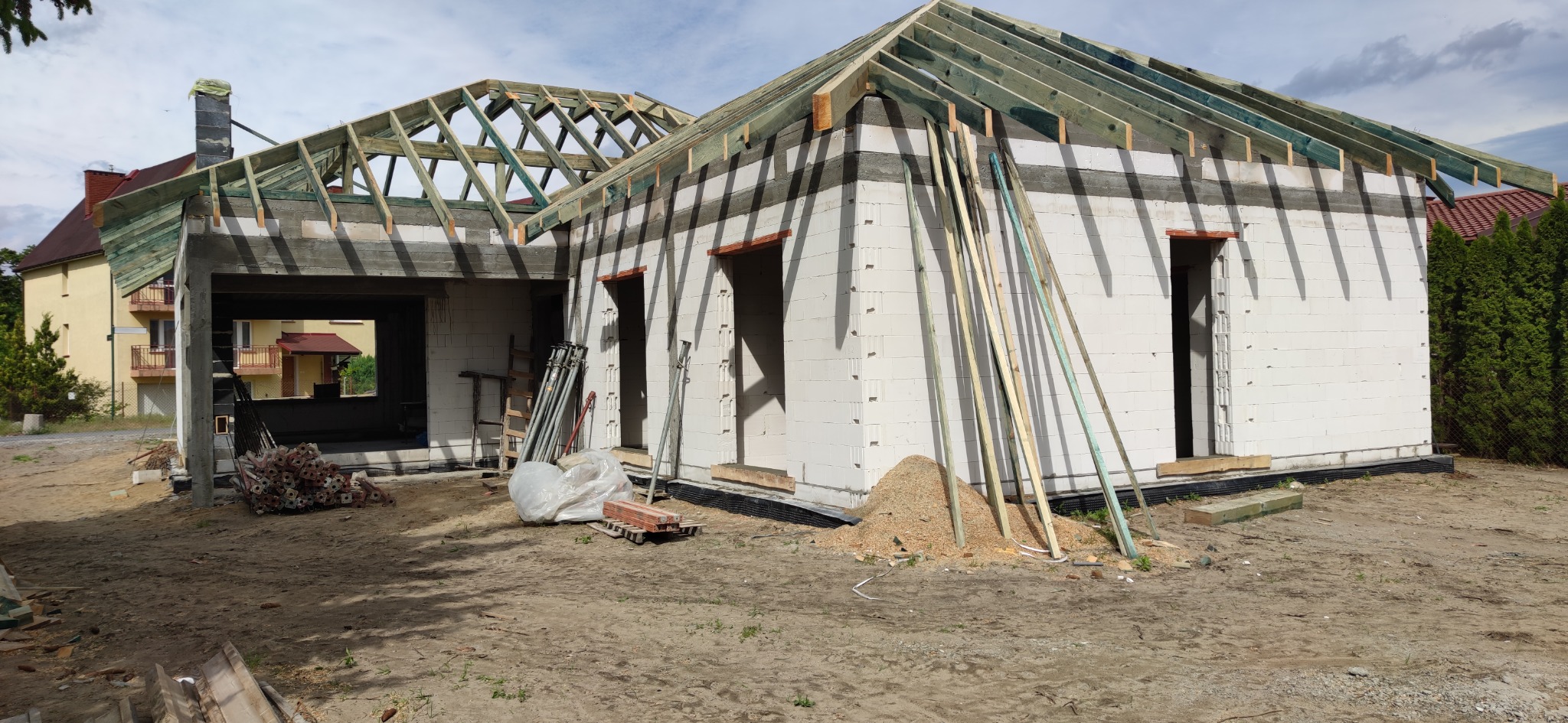 Surowy stan domu murowanego z białych bloczków, widoczne otwory okienne i drzwiowe, drewniana konstrukcja dachu w trakcie montażu, podpory, narzędzia, teren budowy.