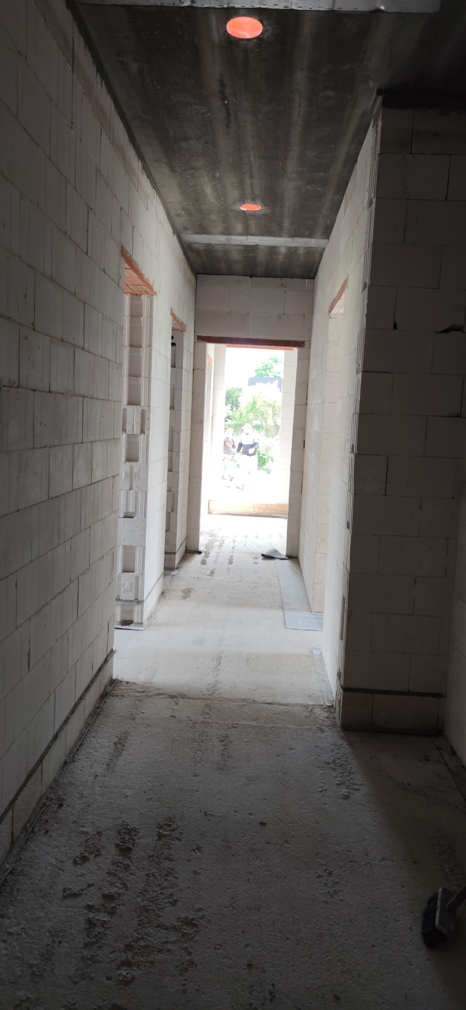 Wnętrze domu w stanie surowym: widok na korytarz z białymi ścianami z betonu komórkowego, surowy betonowy sufit z widocznymi instalacjami elektrycznymi i przygotowanymi otworami na drzwi.