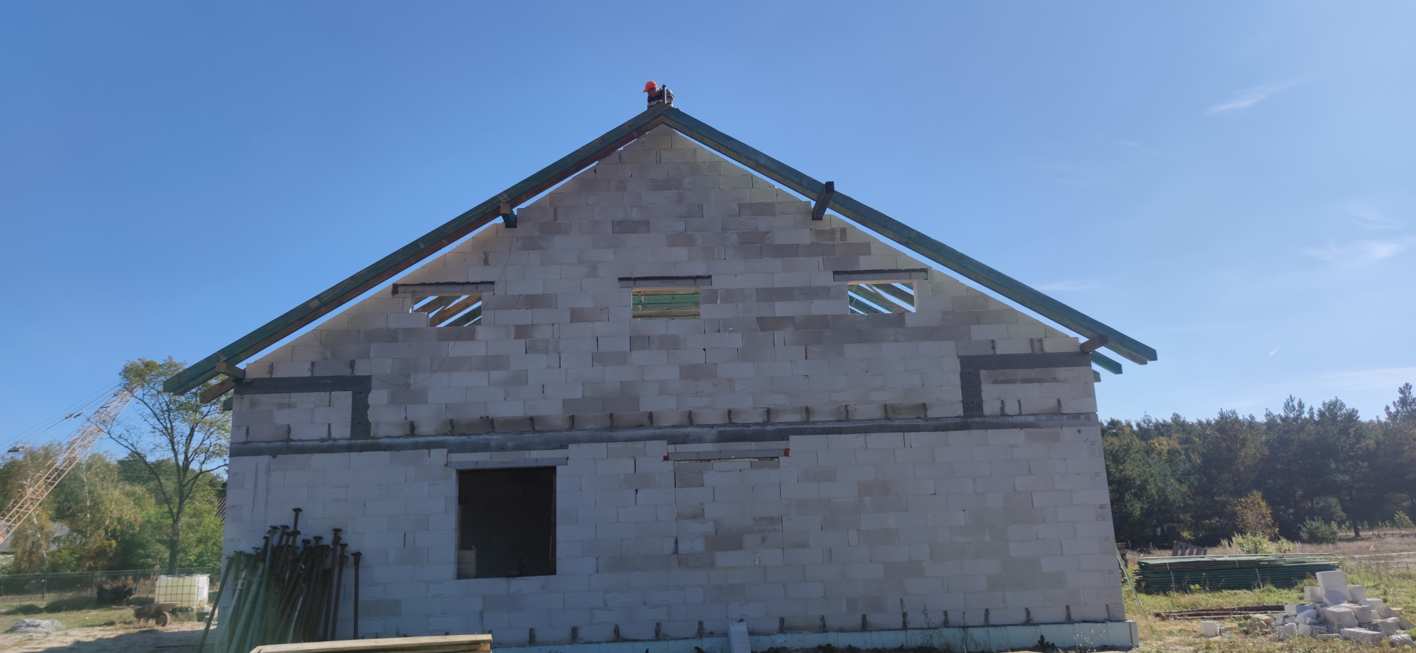 Wznoszenie dwukondygnacyjnego domu z pustaków, widoczne otwory okienne z drewnianymi elementami konstrukcyjnymi oraz częściowo wykonana więźba dachowa, na szczycie dachu robotnik w pomarańczowym...