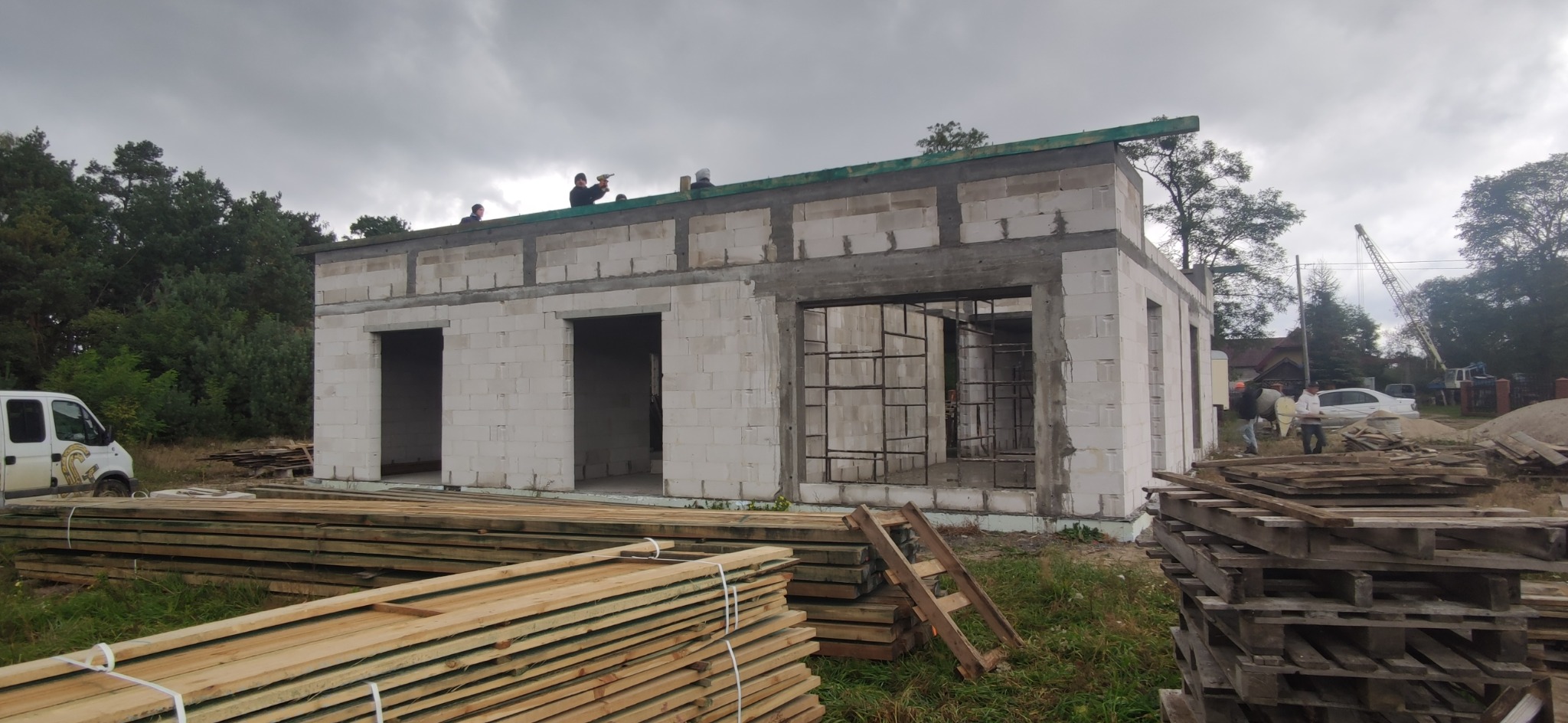 Wznoszenie surowego, parterowego domu z białych bloczków, widoczni pracownicy na dachu, w tle las i pochmurne niebo, na pierwszym planie deski i palety.