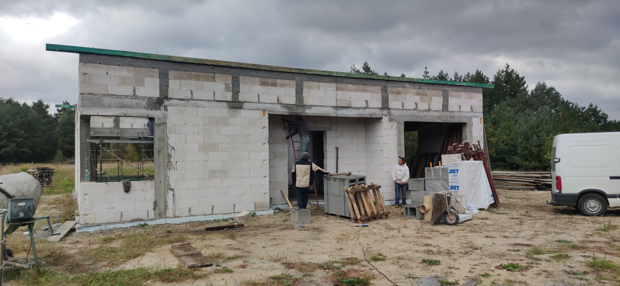 Wznoszenie ścian parterowego budynku z bloczków betonowych; pracownicy na drabinie i obok budynku, widoczny samochód dostawczy i materiały budowlane.