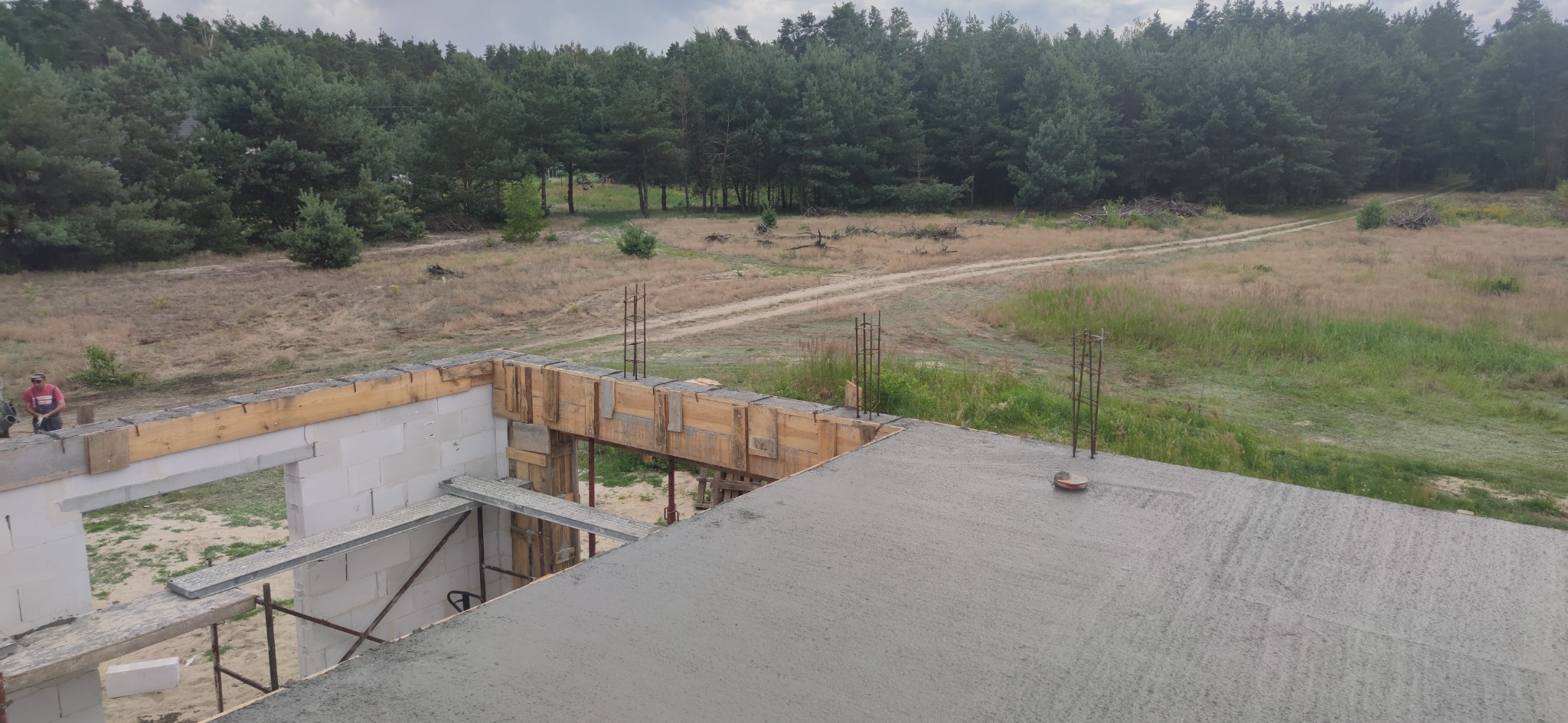 Wylany betonowy strop na budowie domu jednorodzinnego, widoczne szalunki i wystające pręty zbrojeniowe, w tle las i polna droga.