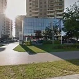 Szklany biurowiec z widocznymi drzewami i wysokimi budynkami mieszkalnymi w tle, oświetlony porannym słońcem.