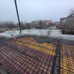 Zalewanie betonem zbrojonej płyty fundamentowej na żółtym szalunku, widok z góry w pochmurny dzień, w tle zabudowania mieszkalne.