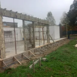 Wznoszenie szkieletu domu z bloczków, widoczne zbrojenie w otworach okiennych i drzwiowych, szalunek fundamentów wzdłuż trawnika, pochmurne niebo w tle.