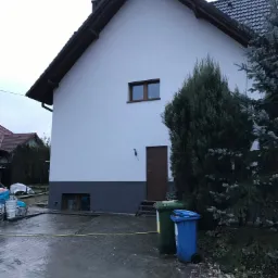 Świeżo otynkowany dom jednorodzinny z białymi ścianami i szarą podmurówką, widoczne dwa kosze na śmieci i pozostałości po pracach budowlanych na podjeździe.