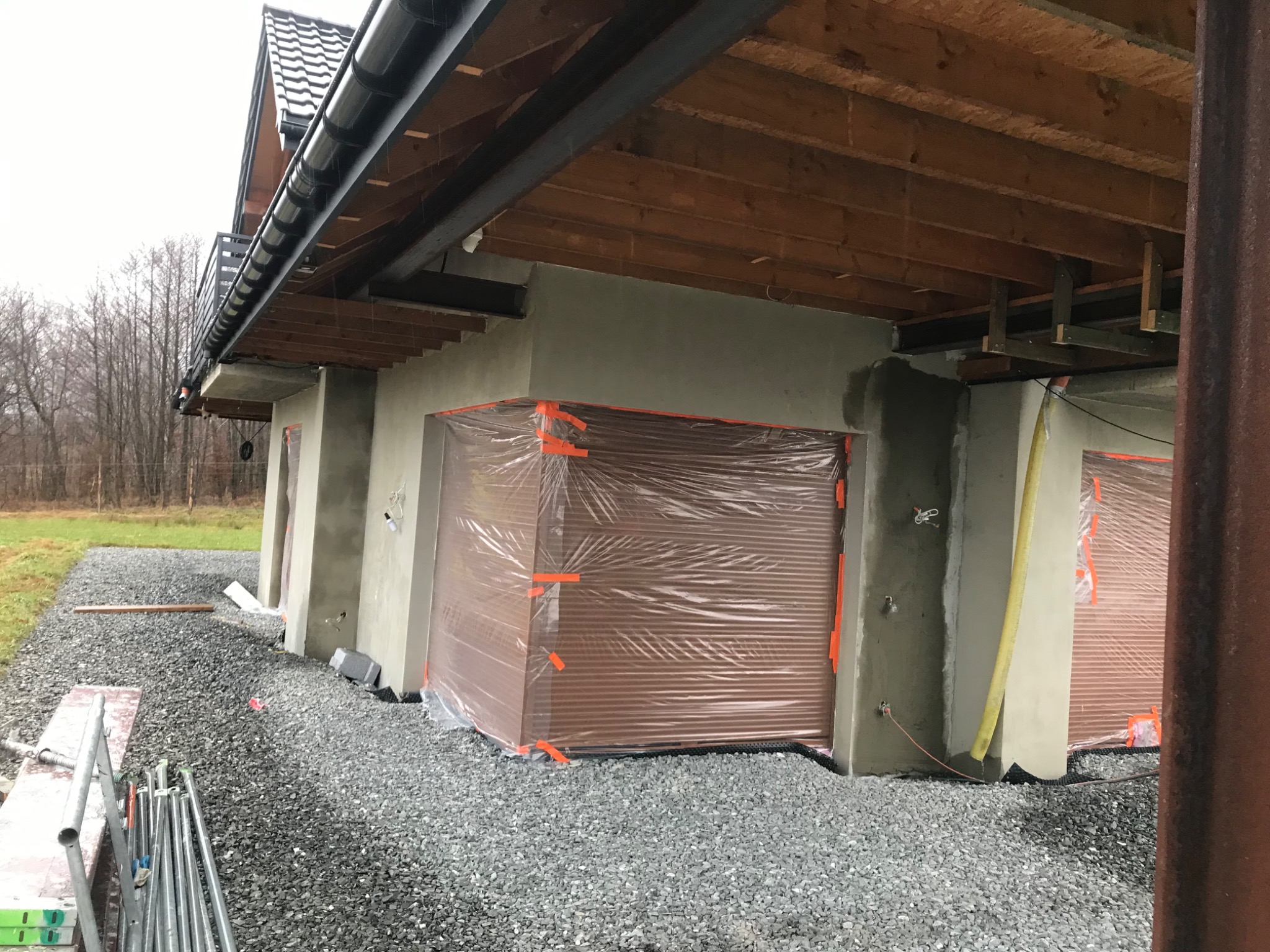 Elewacja budynku w trakcie ocieplania, zabezpieczona folią z pomarańczową taśmą, widoczne rusztowania i teren wokół budynku wysypany żwirem.