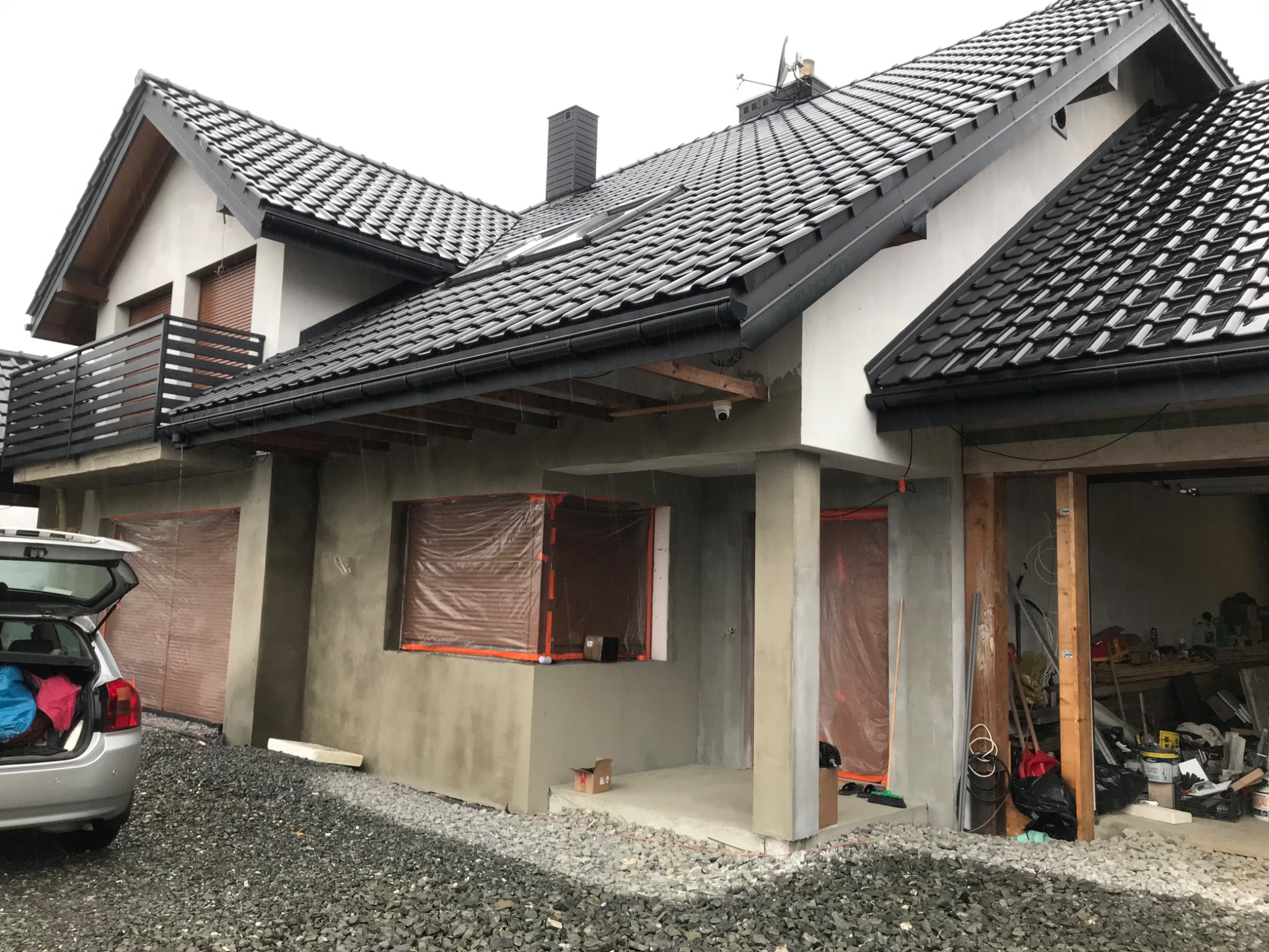 Dom w trakcie prac elewacyjnych, z widocznym świeżo nałożonym tynkiem, zabezpieczonymi oknami folią i otwartym bagażnikiem samochodu osobowego. W tle czarny dach z dachówki ceramicznej.