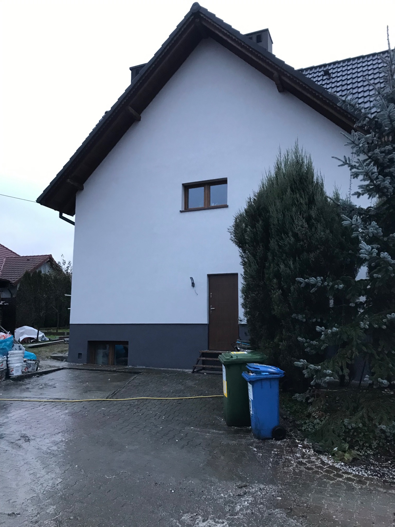 Świeżo otynkowany dom jednorodzinny z białymi ścianami i szarą podmurówką, widoczne dwa kosze na śmieci i pozostałości po pracach budowlanych na podjeździe.