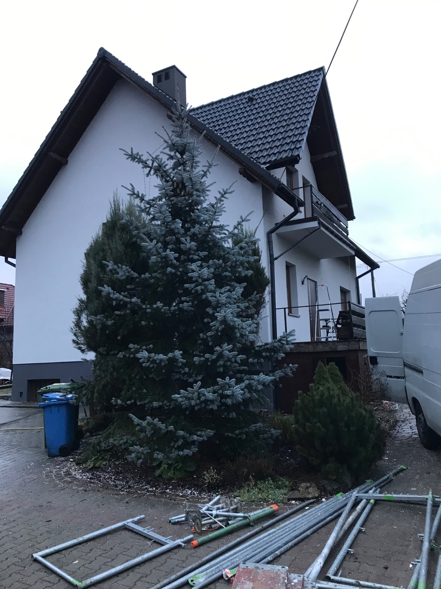 Biały dom jednorodzinny z balkonem, czarnym dachem i dużą choinką przed elewacją, widoczne rusztowania i samochód dostawczy.
