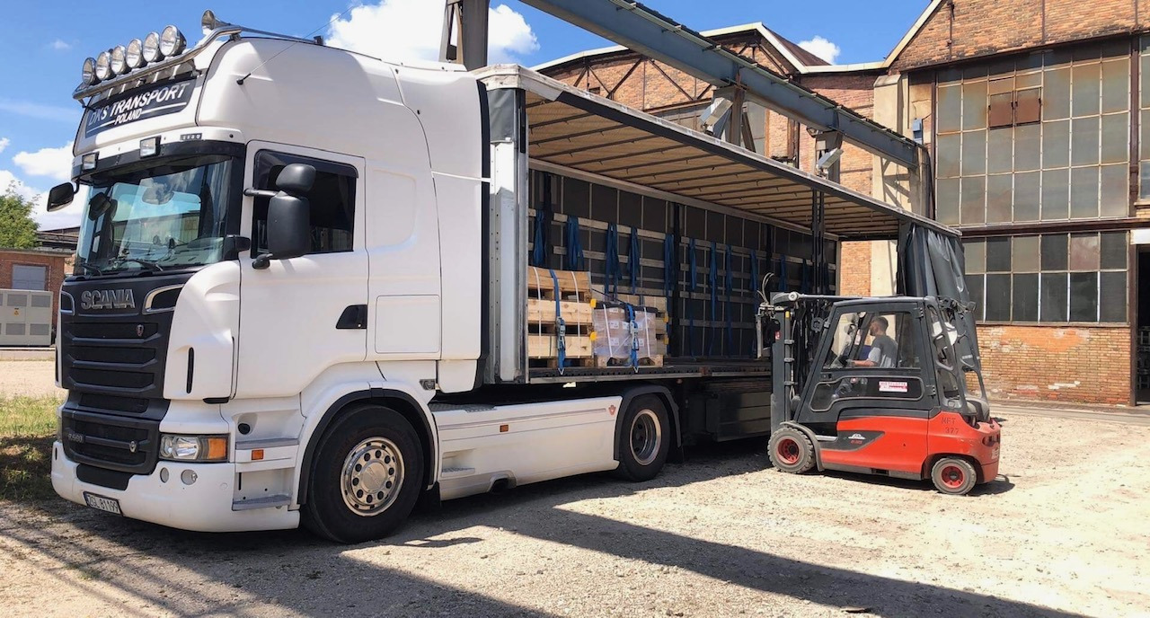 Biała ciężarówka Scania z otwartą naczepą, do której podjeżdża pomarańczowy wózek widłowy z operatorem, na paletach ładunek zabezpieczony niebieskimi pasami transportowymi, w tle ceglany budynek...