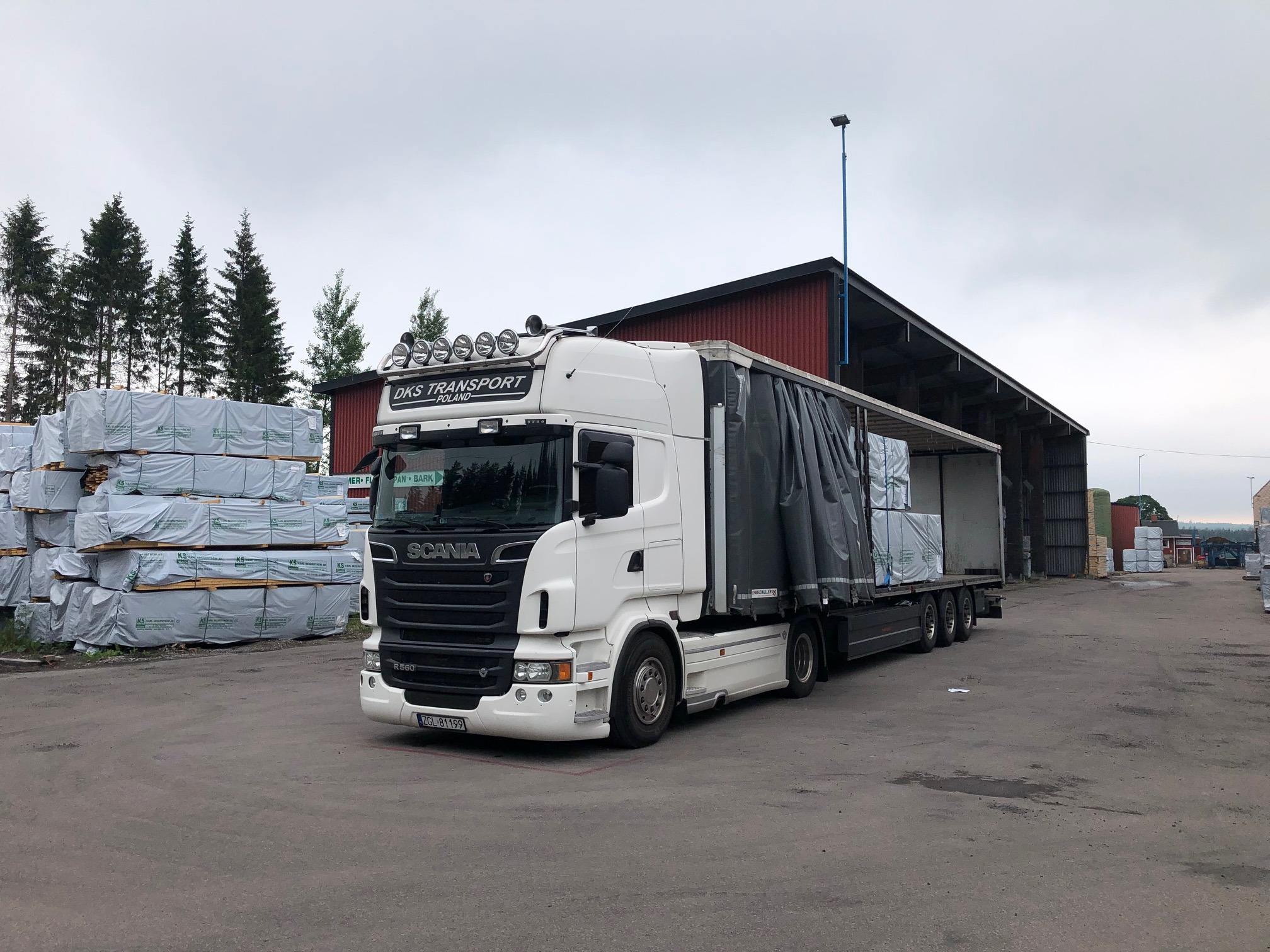 Biała ciężarówka Scania z naczepą, częściowo załadowana drewnem, na placu składowym z widocznymi stertami zapakowanego drewna w tle i budynkiem magazynowym w pochmurny dzień.