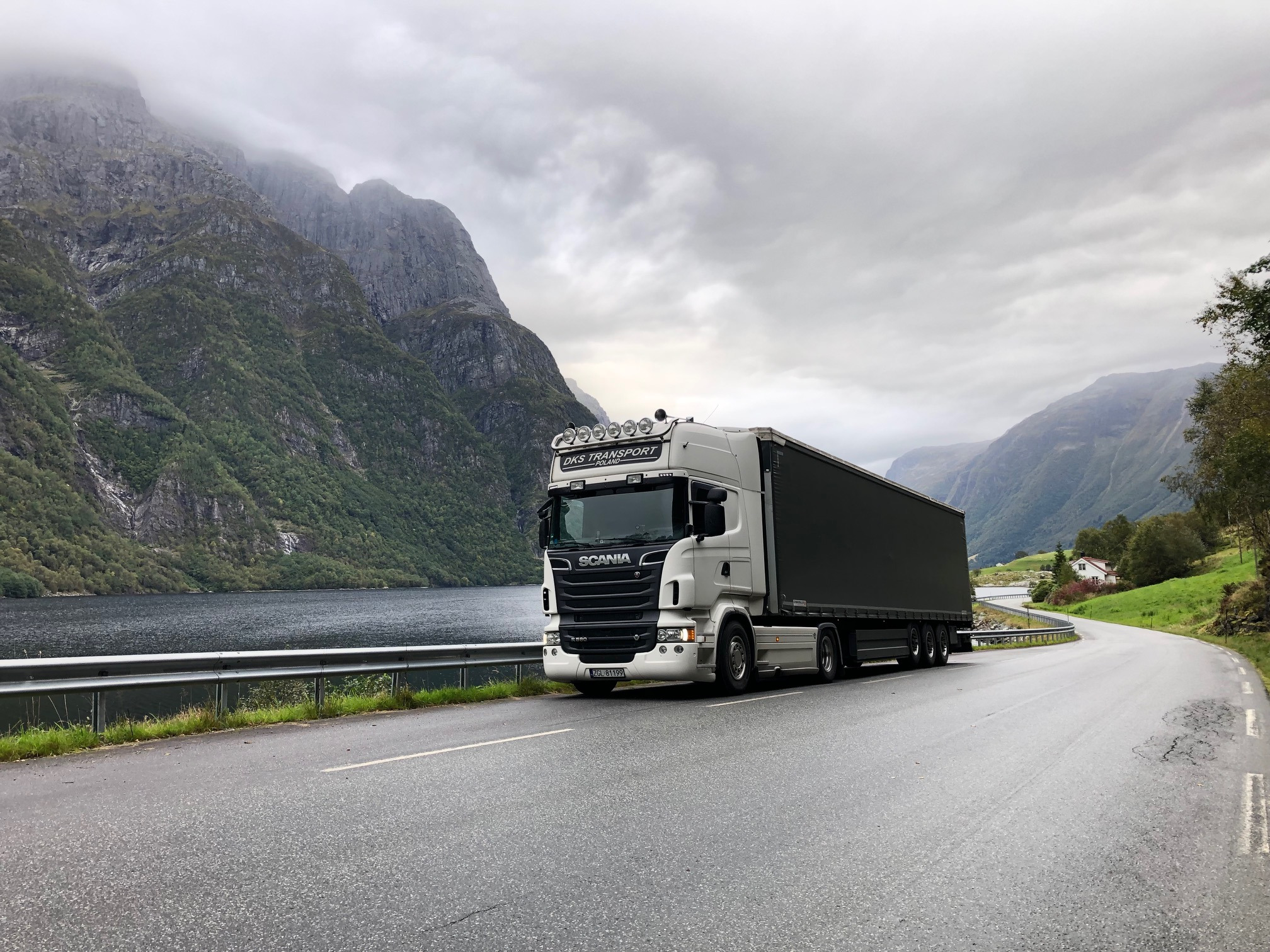 Biała ciężarówka Scania z czarną naczepą zaparkowana na mokrej drodze z widokiem na fiord i góry we mgle.