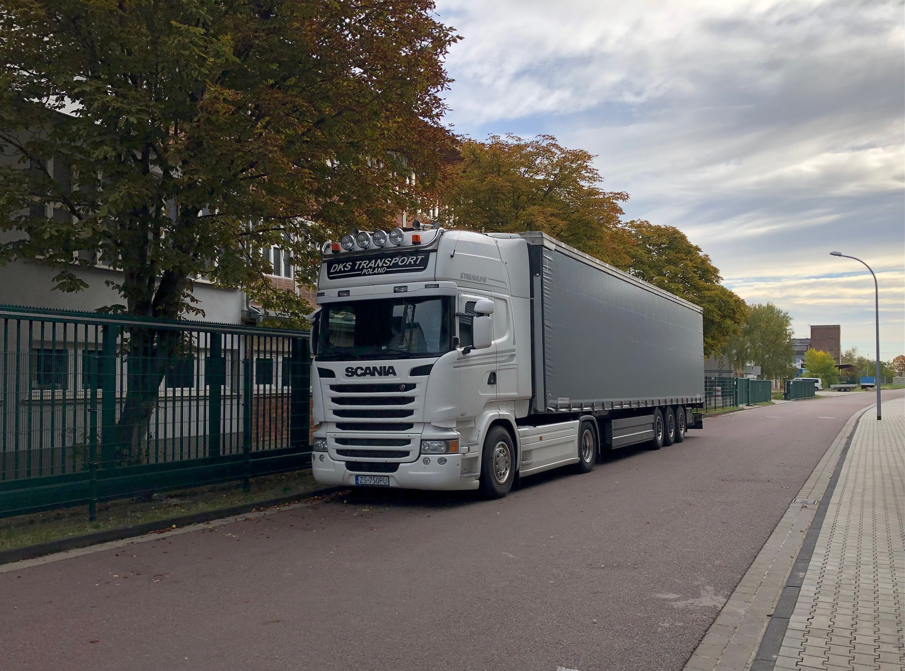 Biała ciężarówka Scania z szarą naczepą zaparkowana na ulicy w jesiennej scenerii, widoczny budynek z zielonym ogrodzeniem w tle.