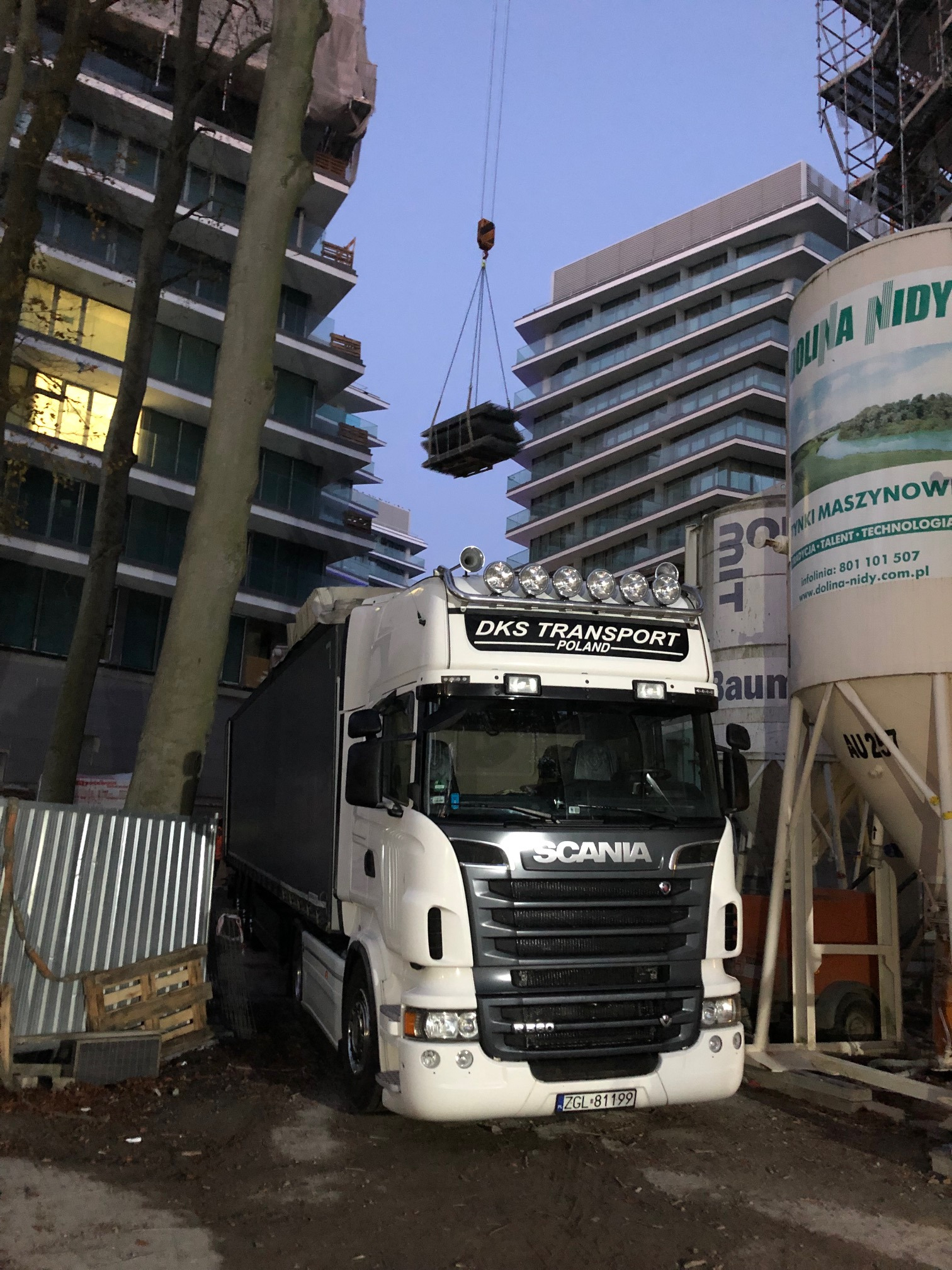 Biała ciężarówka Scania firmy DKS Transport Poland na tle budowy, z dźwigiem podnoszącym ładunek i silosami Dolina Nidy.