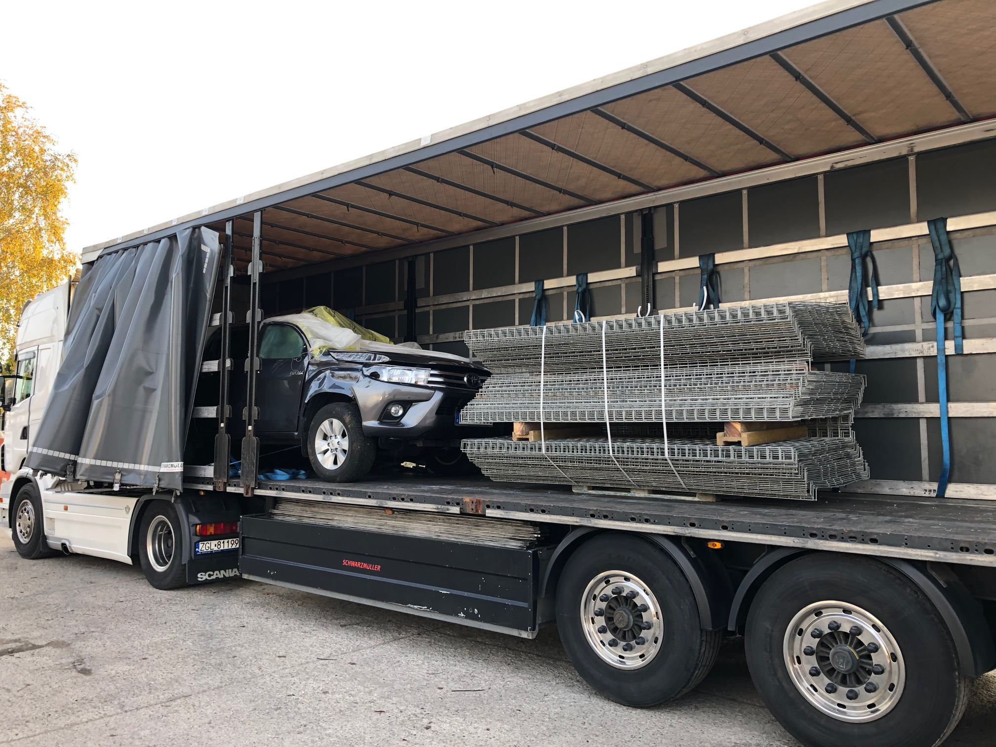 Ciężarówka Scania z otwartą plandeką przewozi szarą Toyotę Hilux przykrytą folią i paletę z siatkami ogrodzeniowymi zabezpieczonymi pasami.