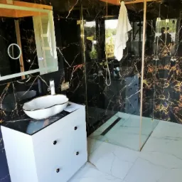 Elegancka łazienka z czarnymi marmurowymi ścianami, białą umywalką na szafce z trzema szufladami i kabiną prysznicową ze złotymi detalami.