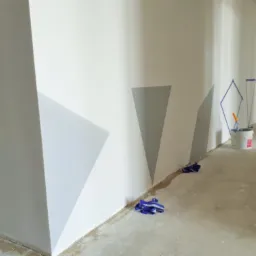 Wnętrze w trakcie malowania z geometrycznymi wzorami w odcieniach bieli i szarości, zabezpieczone taśmą malarską, widoczny kubeł z farbą i szmatki na podłodze.