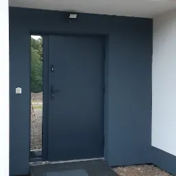 Wejście do budynku z ciemnoszarą elewacją i drzwiami, widoczny próg i kamieniste podłoże.