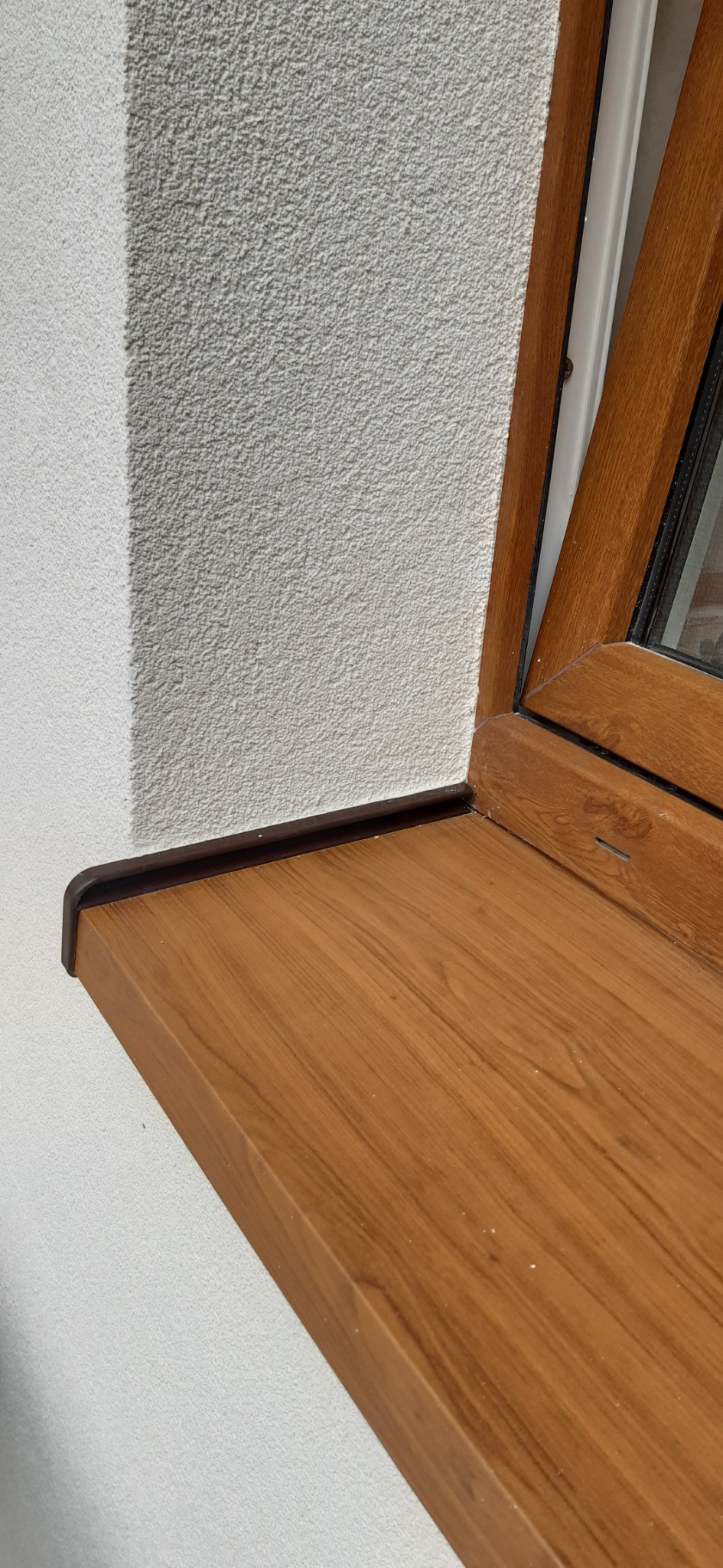 Detale wykończenia okna z drewnopodobnym parapetem zewnętrznym i jasną elewacją o wyraźnej strukturze.