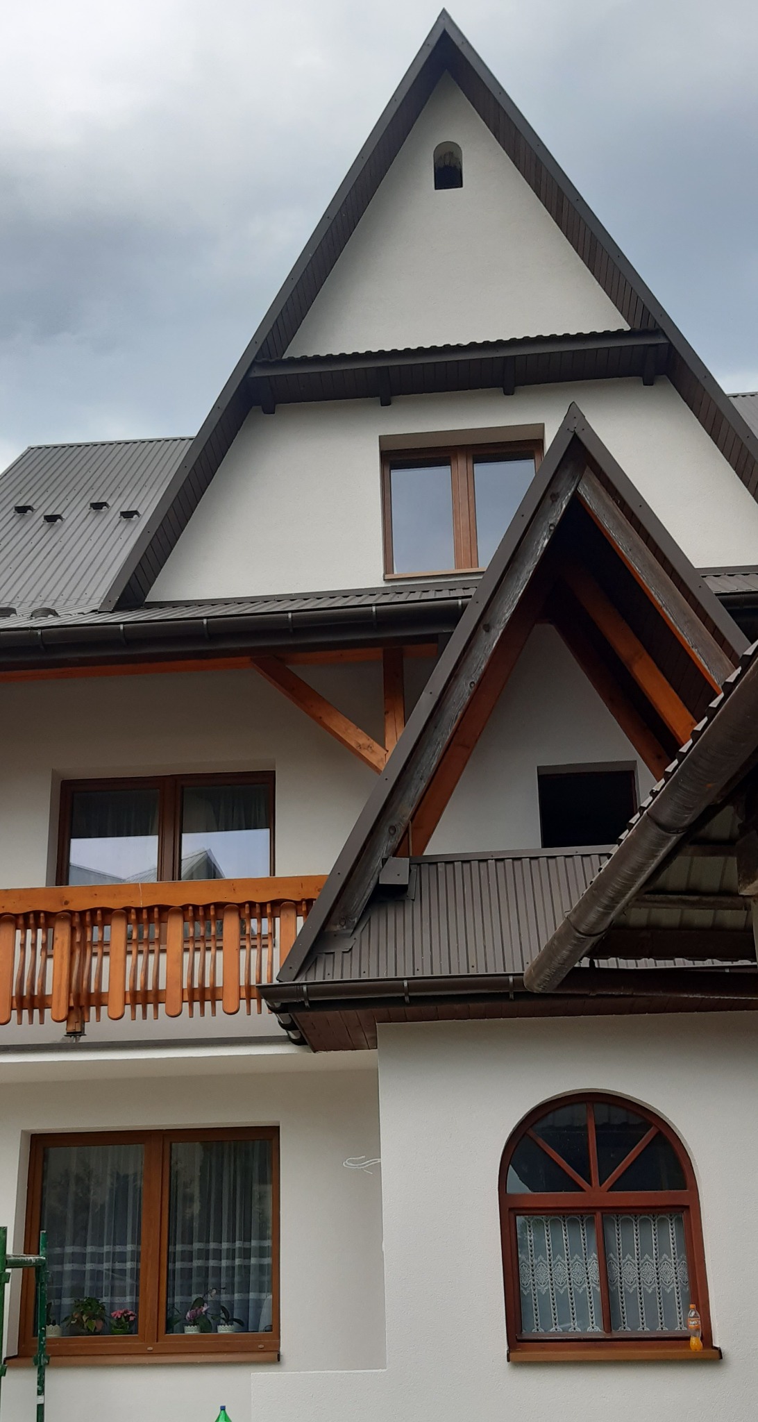 Fragment białej elewacji domu w Zabrzeżu z drewnianymi elementami, balkonem i oknami o różnych kształtach