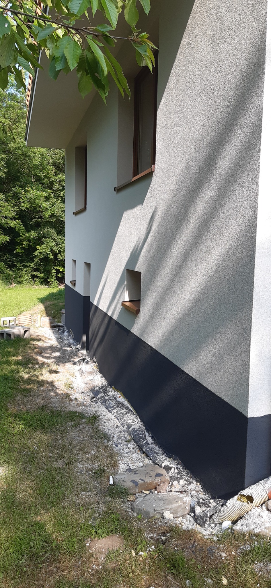 Fragment elewacji domu z szarą, teksturowaną górną częścią i ciemnoszarą dolną częścią, widoczne małe okienka z drewnianymi parapetami, cień drzewa na ścianie, oraz resztki materiałów budowlanych...