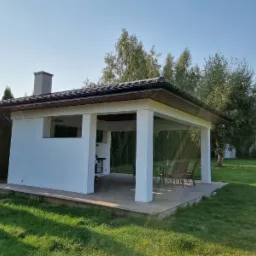 Biała murowana altana z kominem i zadaszeniem, wyposażona w grill i stół z krzesłami, otoczona zielonym trawnikiem i drzewami.