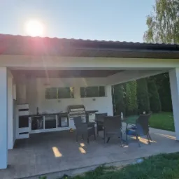 Zadaszona, letnia kuchnia z grillem gazowym, blatem roboczym i szafkami, otoczona zielenią, z widocznymi elementami wykończenia i meblami ogrodowymi.