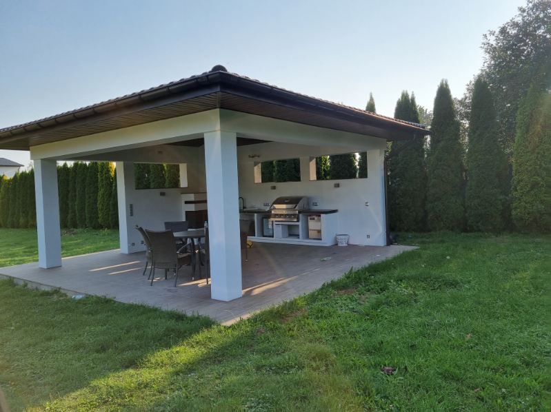 Nowoczesna altana ogrodowa z grillem, stołem i krzesłami, wykończona w bieli i brązie, otoczona zielonym trawnikiem i tujami.