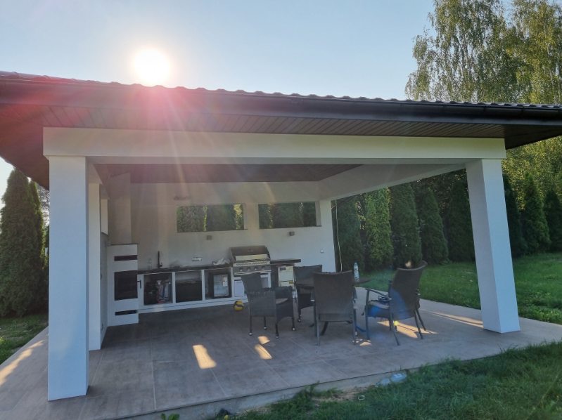 Zadaszona, letnia kuchnia z grillem gazowym, blatem roboczym i szafkami, otoczona zielenią, z widocznymi elementami wykończenia i meblami ogrodowymi.