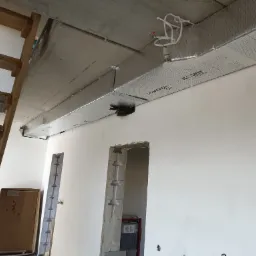 Montaż izolowanego kanału wentylacyjnego Climaver Isover 360 pod betonowym sufitem w surowym wnętrzu, widoczne fragmenty drewnianych schodów i niedokończone otwory w ścianie.