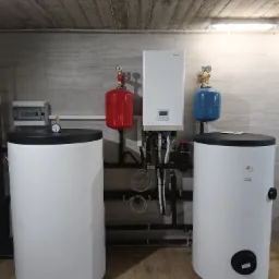 Zestaw instalacji grzewczej w kotłowni: biały piec z wyświetlaczem, dwa białe zbiorniki z czarnymi pokrywami, czerwony i niebieski zbiornik wyrównawczy, orurowanie, szara ściana z płytkami...