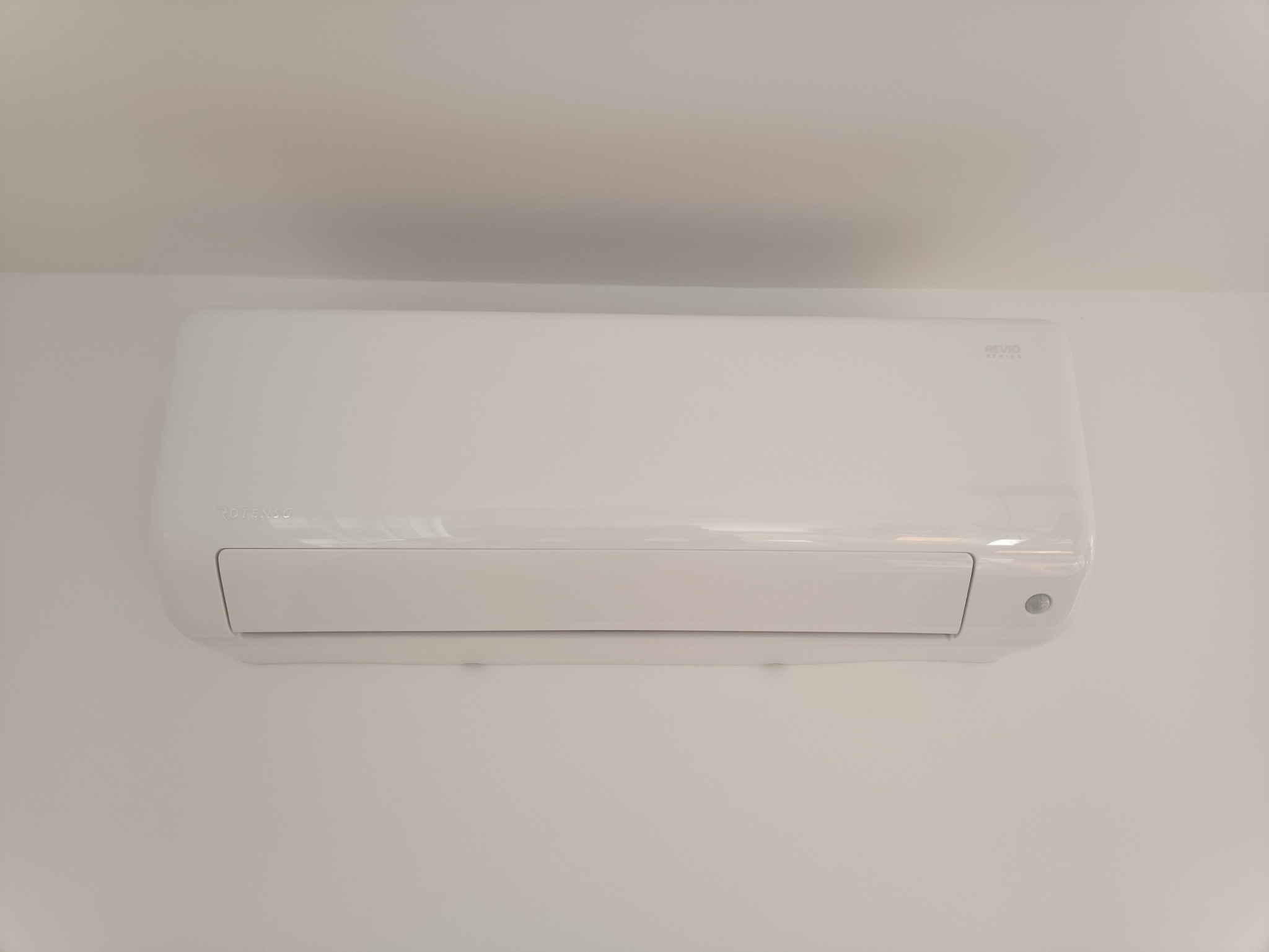 Biała jednostka wewnętrzna klimatyzatora marki Rotenso Revio Series zamontowana na białej ścianie, widok centralny.