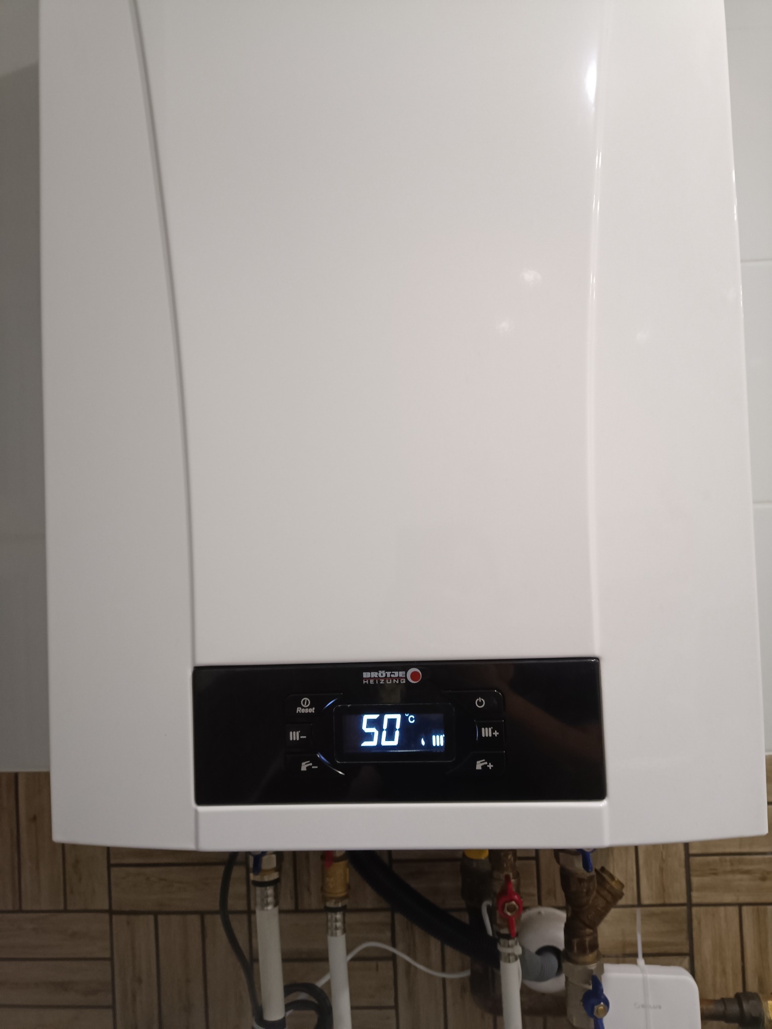 Biały, wiszący piec gazowy marki Brötje z wyświetlaczem pokazującym temperaturę 50 stopni Celsjusza, zamontowany na ścianie wyłożonej płytkami imitującymi drewno, z widocznymi rurami i zaworami.
