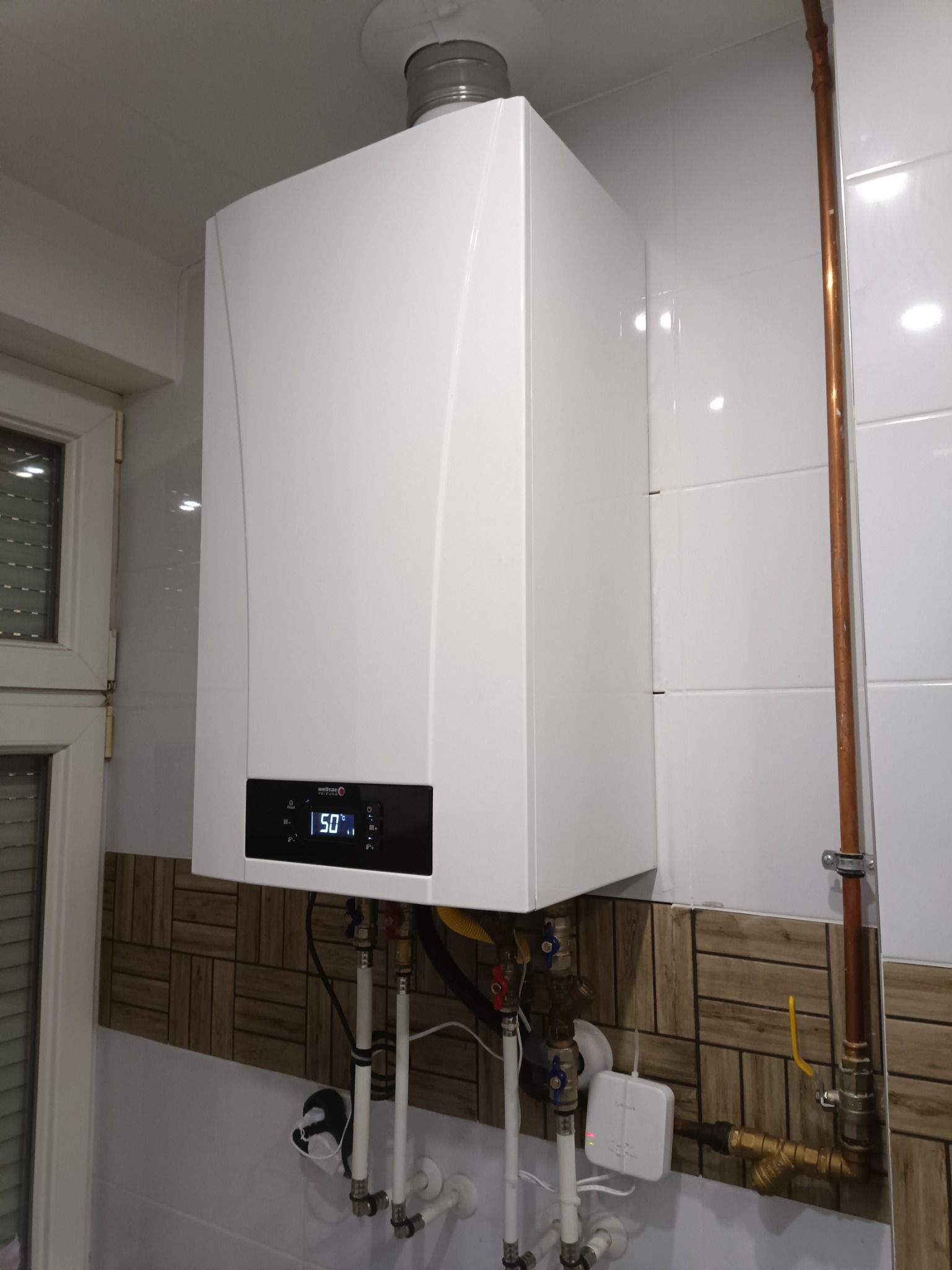 Biały, nowoczesny piec gazowy zamontowany na ścianie z białych i brązowych płytek, widoczne rury doprowadzające media, na wyświetlaczu temperatura 50 stopni Celsjusza.