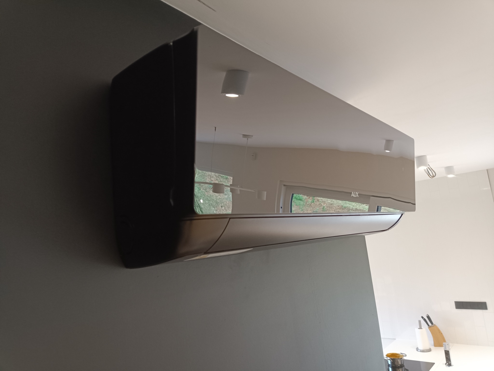 Czarny klimatyzator AUX zamontowany na szarej ścianie w nowoczesnym wnętrzu, widoczne odbicie okna i oświetlenia sufitowego.