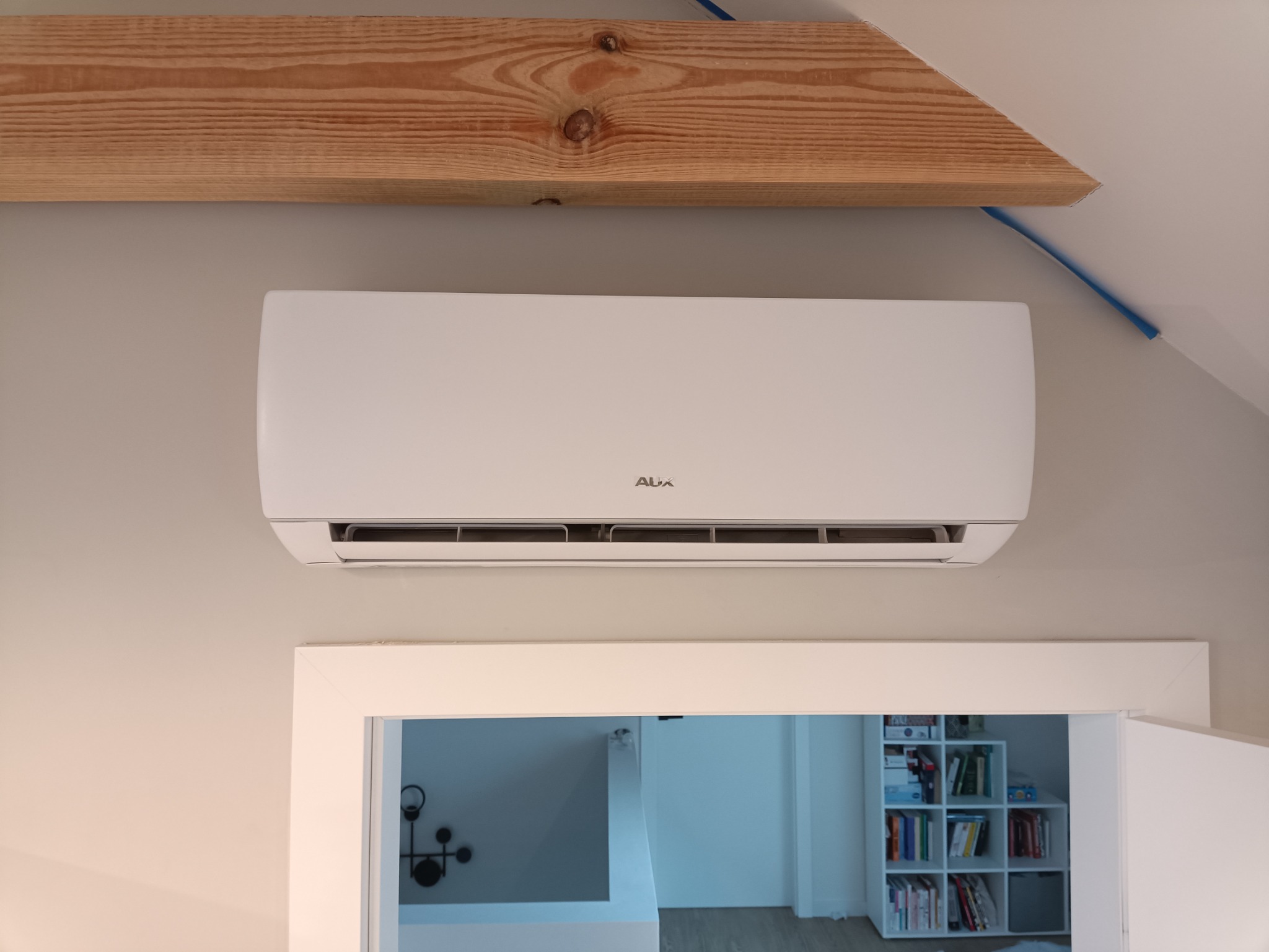Biały klimatyzator marki AUX zamontowany na ścianie nad białymi drzwiami, widoczna drewniana belka stropowa i fragment instalacji elektrycznej.