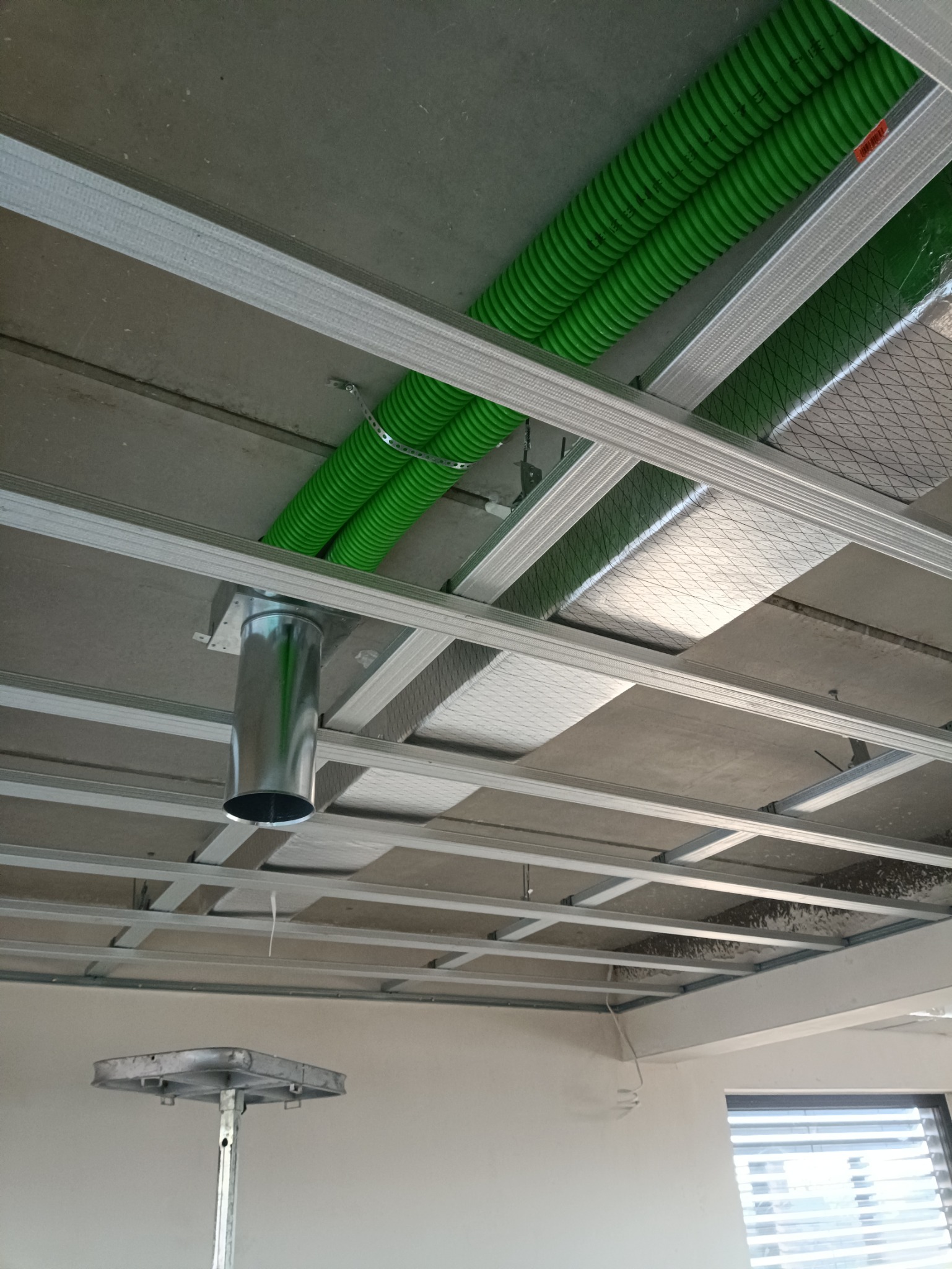 Instalacja podwieszanego sufitu z widocznymi zielonymi rurami wentylacyjnymi i metalowymi kanałami, w trakcie montażu.
