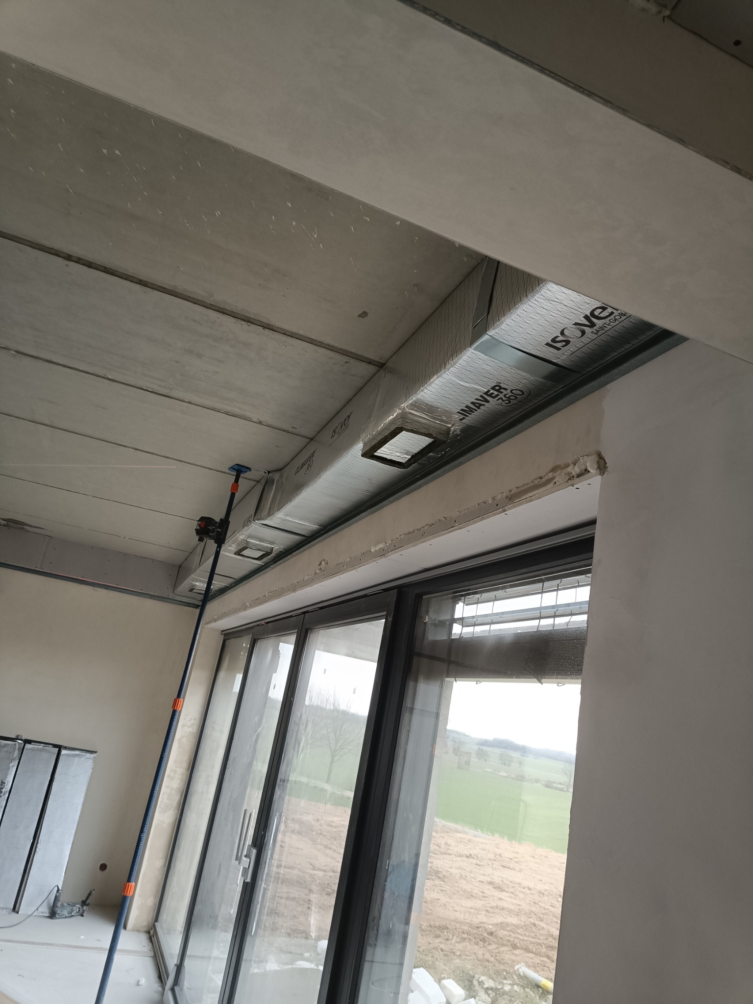 Montaż kanału wentylacyjnego Isover ClimAver 360 z kratkami w nowym budynku, widok z dołu na sufit podwieszany i okno z widokiem na pole.