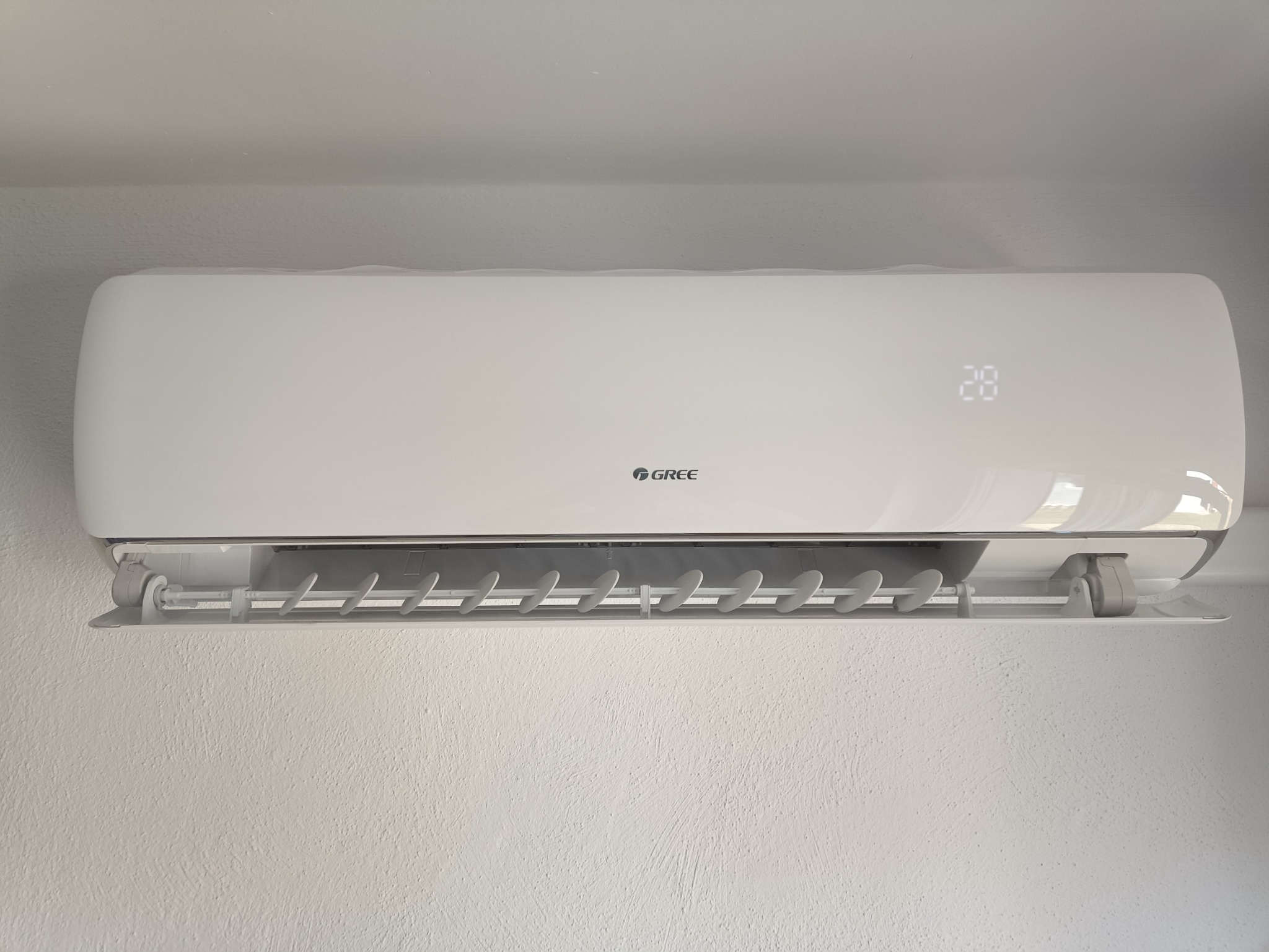 Biały klimatyzator marki Gree zamontowany na jasnej, teksturowanej ścianie, wyświetlający temperaturę 28 stopni Celsjusza.