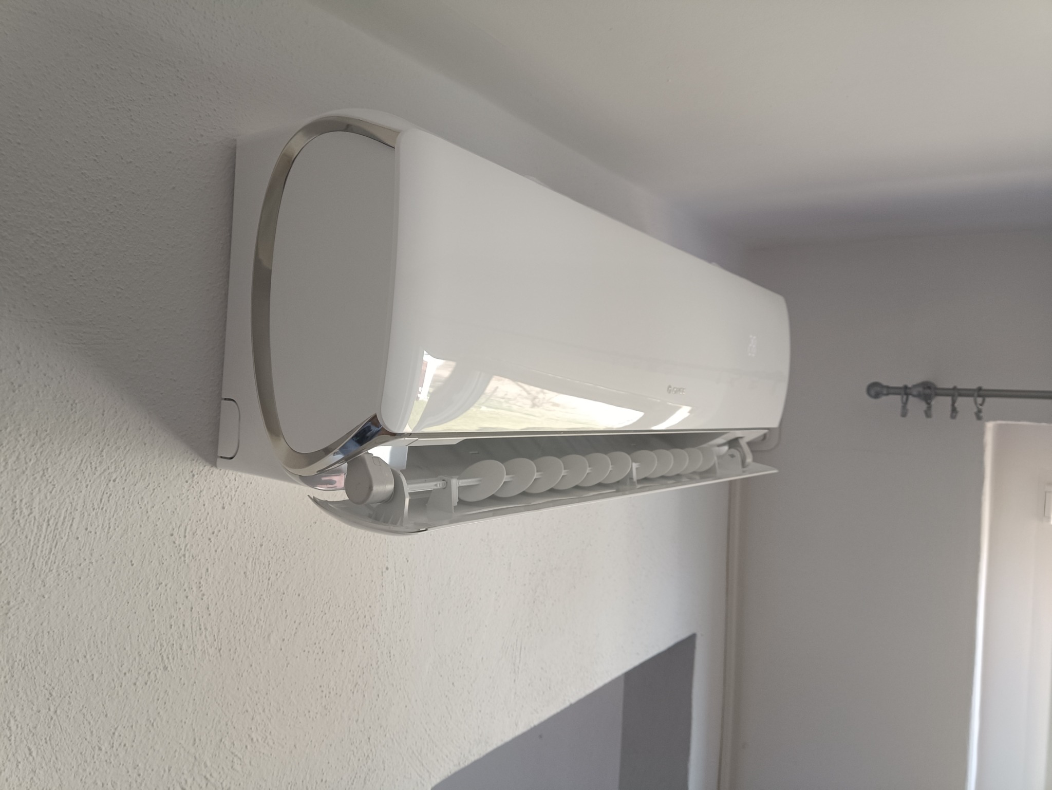 Nowoczesny, biały klimatyzator z chromowanymi akcentami, zamontowany na teksturowanej ścianie w jasnym wnętrzu, widoczne żaluzje nawiewu powietrza.