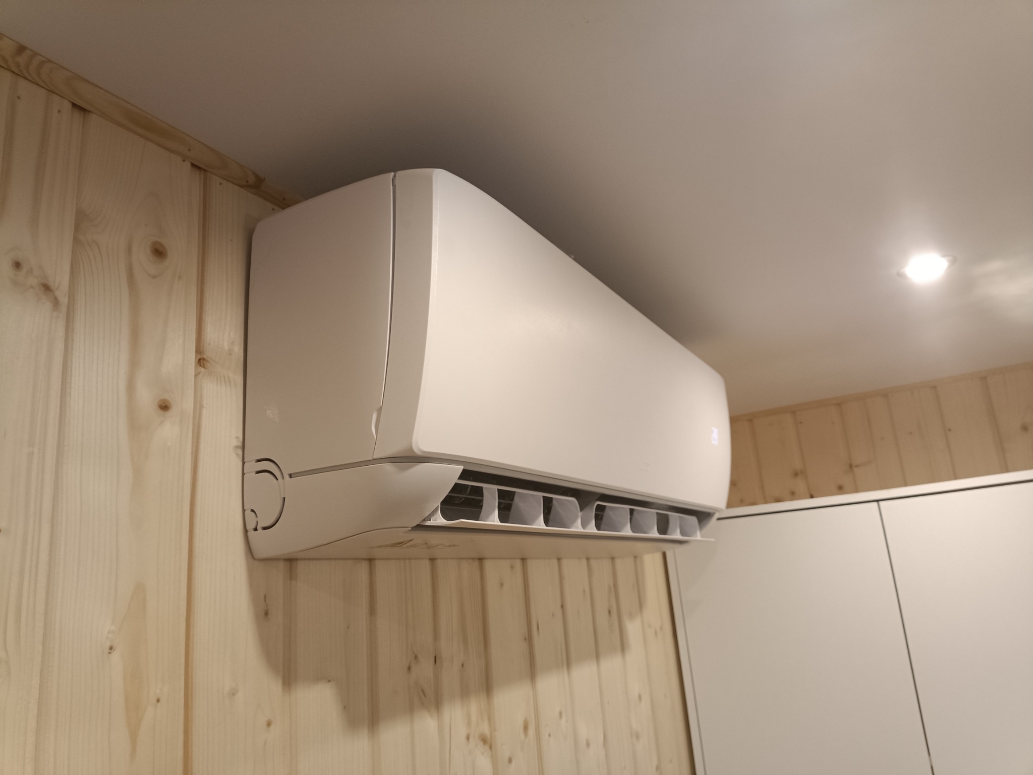 Biała jednostka wewnętrzna klimatyzatora zamontowana na ścianie z boazerii w jasnym odcieniu drewna, widok z boku, w tle białe szafki i oświetlenie sufitowe.