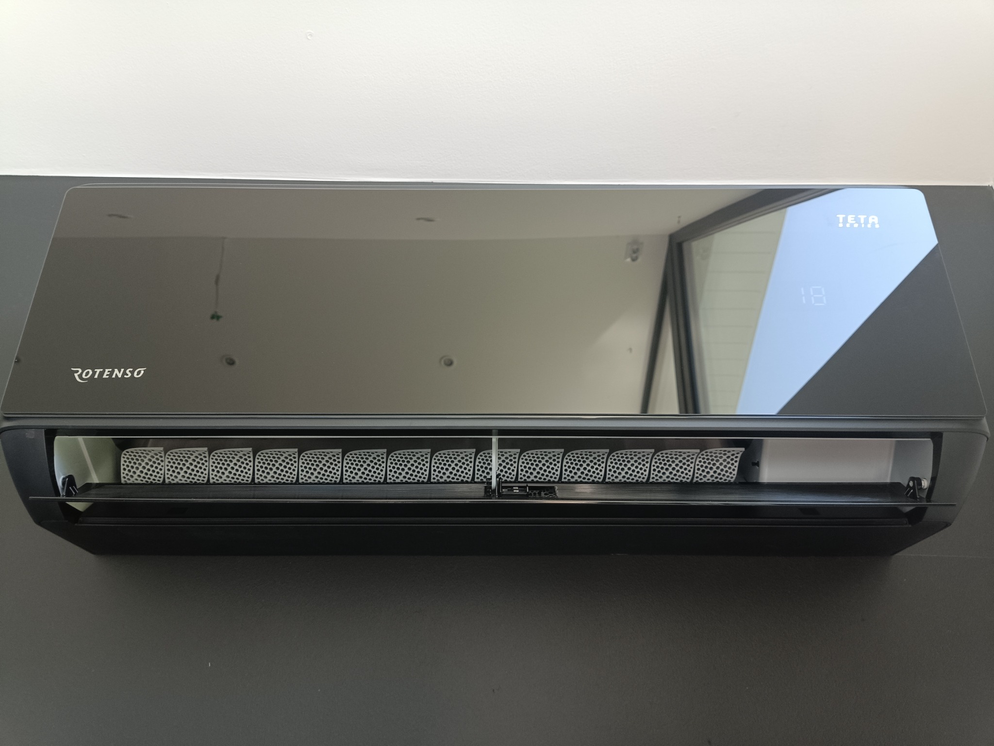 Czarny klimatyzator Rotenso Teta Series zamontowany na szarej ścianie, widoczny wyświetlacz temperatury.