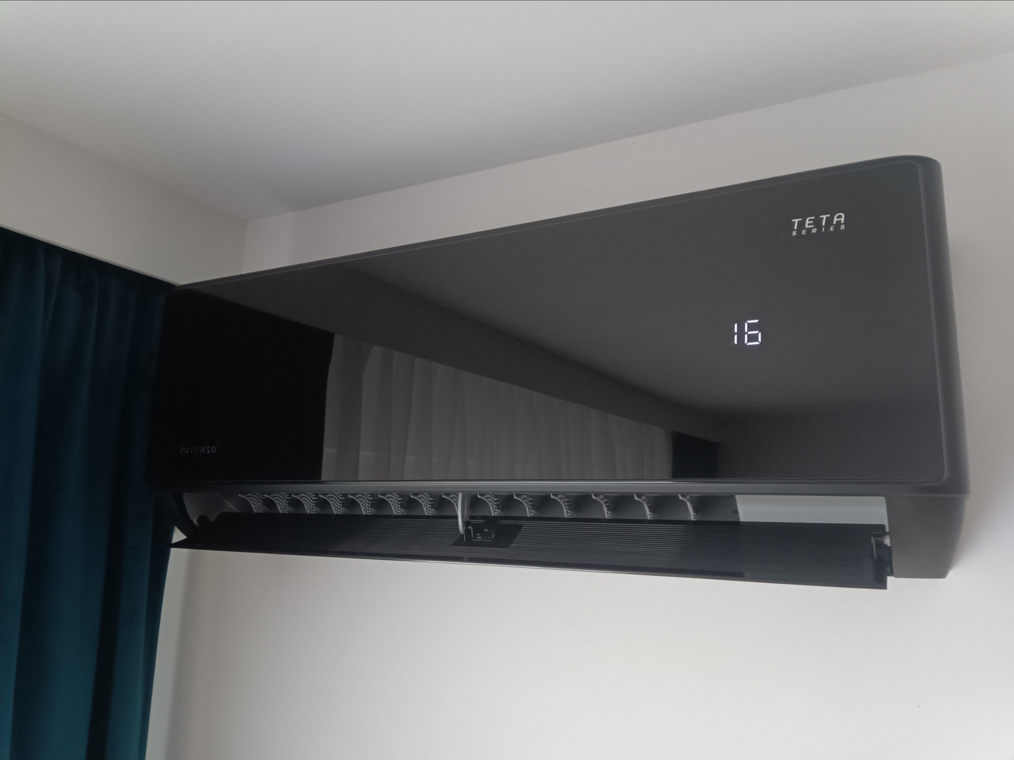 Czarny klimatyzator TETA Series zamontowany na białej ścianie, wyświetlający temperaturę 16 stopni, z widoczną zasłoną w tle.