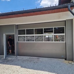 Mariobramy Mariusz Byczek - Szara brama garażowa segmentowa z oknami, widoczny pracownik w wejściu bocznym, żwirowe podłoże.