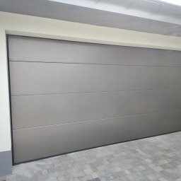 Mariobramy Mariusz Byczek - Zamknięta, segmentowa brama garażowa w kolorze szarym, z poziomymi przetłoczeniami, zamontowana w garażu w Rybniku. Widoczny fragment elewacji budynku i kostka brukowa przed garażem.