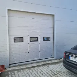 Szara segmentowa brama z oknami i drzwiami wejściowymi w budynku przemysłowym, widoczny fragment zaparkowanego samochodu osobowego.