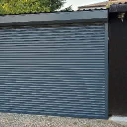 Zewnętrzna, szara roleta na budynku gospodarczym, widoczny fragment ogrodu i drzewo w tle.