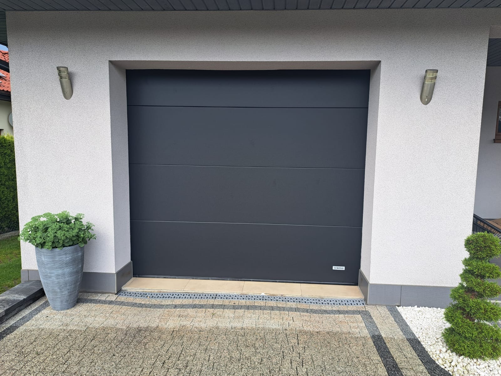 Nowoczesna, antracytowa brama garażowa segmentowa z poziomymi przetłoczeniami, z widocznym logo producenta w prawym dolnym rogu, zamontowana w jasnym garażu z oświetleniem zewnętrznym.